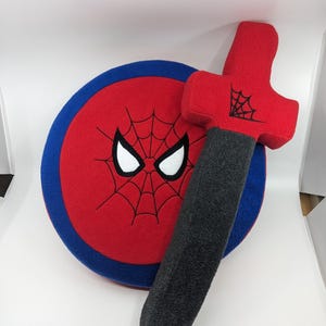 May include: A red and blue plush toy shield with a black embroidered spider-man face and web design. A gray and red plush toy sword with a black embroidered web design.