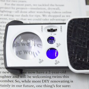 May include: A white and black handheld magnifying glass with a built-in UV light. The magnifying glass has a lens with a 30x, 60x, and 90x magnification. The text "LED90X" and "LED60X" is printed on the side of the magnifying glass.