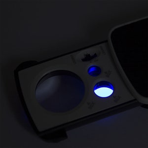 May include: A white and black handheld magnifying glass with a blue LED light. The magnifying glass has a round lens and a handle with buttons labeled "LED", "UV", "30X", "60X", and "100X".