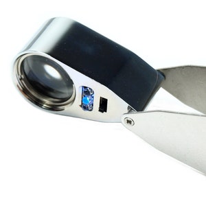 May include: A silver and black magnifying glass with a built-in LED light. The magnifying glass has a folding handle and a clear lens.