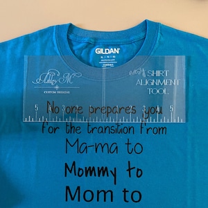 May include: A turquoise Gildan t-shirt with the text "No one prepares you for the transition from Ma-ma to Mommy to Mom to BRUH" in black. A clear shirt alignment tool is placed on the shirt.
