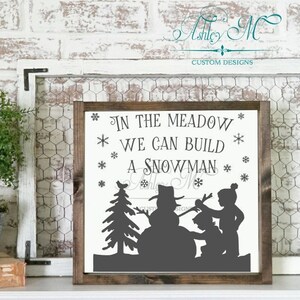 May include: A white wooden sign with the text "In the meadow we can build a snowman" in black lettering. The sign features a silhouette of two children building a snowman in front of a pine tree.