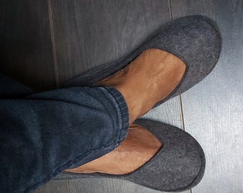 Può includere: Un paio di pantofole in feltro grigio con punta arrotondata e un design semplice. Le pantofole sono indossate ai piedi.
