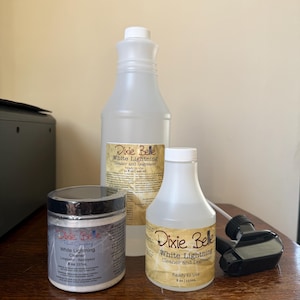 May include: Three Dixie Belle White Lightning cleaning products are displayed on a wooden surface. The products include a jar, a large bottle, and a smaller bottle with a spray nozzle. The labels feature the product name and brand.