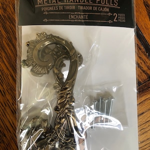 May include: A pair of ornate gold metal drawer pulls with a scroll design. The package includes two pulls and screws. The package says "Kacha Metal Handle Pulls Enchanté 2 Pieces"