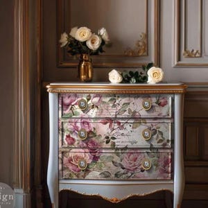 May include: A three-drawer chest with a floral design featuring pink and purple roses. The chest has gold accents, ornate drawer pulls, and a white and light blue painted finish. A vase of white roses sits on top.