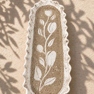 May include: An elongated, oval-shaped ceramic tray with a ruffled, scalloped edge. The tray features a speckled, light-colored rim and a textured, tan-colored center decorated with a white, hand-painted botanical illustration of a leafy stem.