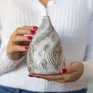 May include: A white ceramic vase with a textured, leaf-like pattern. The vase has a teardrop shape and a narrow neck. The design features carved lines and a contrasting brown interior, creating a unique decorative piece.
