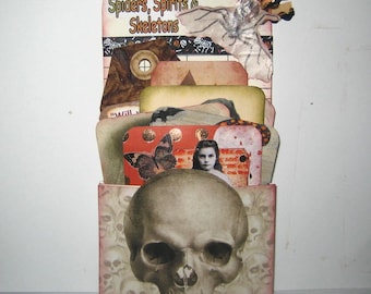 May include: A Halloween-themed decorative box with a skull on the front. The box is filled with layered cards featuring images of a butterfly, a woman, and other spooky elements. The top of the box reads "Spiders, Spirits & Skeletons".