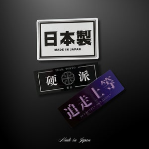 Op de afbeelding: Drie rechthoekige stickers met Japanse tekst en Engelse vertalingen. De bovenste sticker is wit met zwarte tekst met de tekst "Made in Japan" en "日本製". De middelste sticker is zwart met witte tekst. De onderste sticker is paars met witte tekst.