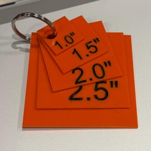 May include: A set of four square orange acrylic tags stacked on a metal keyring. Each tag features black printed text indicating its size: 1.0 inch, 1.5 inches, 2.0 inches, and 2.5 inches. The tags are arranged in ascending order of size.
