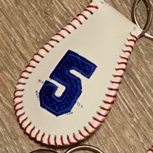May include: White baseball-style keychain with red stitching and a blue felt number 5. The keychain has a silver metal key ring.