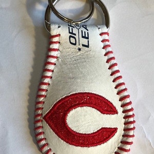 May include: White leather baseball keychain with red stitching and a red embroidered "C" on the front. The keychain has a silver metal key ring and says "Official League" on the back.