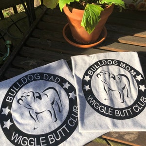 May include: Two grey t-shirts with black text and a black silhouette of a bulldog. The text on the shirts reads "Bulldog Dad Wiggle Butt Club" and "Bulldog Mom Wiggle Butt Club".