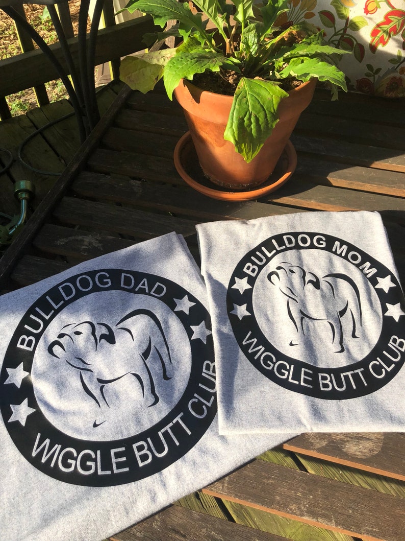 May include: Two grey t-shirts with black text and a black silhouette of a bulldog. The text on the shirts reads "Bulldog Dad Wiggle Butt Club" and "Bulldog Mom Wiggle Butt Club".