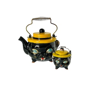 May include: A black ceramic teapot and sugar bowl set, both shaped like cat faces. The teapot has a yellow lid and handle, and the sugar bowl has a matching lid. The cat faces have blue eyes and red mouths. The teapot has a metal handle.