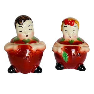 May include: A pair of vintage ceramic cookie jars shaped like apples. The jars are decorated with painted faces of a boy and a girl. The boy has brown hair and the girl has blonde hair. Both jars have green leaves and red accents.