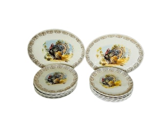 May include: A collection of vintage ceramic plates and platters with a turkey motif. The set comprises two oval serving platters and two stacks of smaller plates. The plates and platters have a cream base with gold edging.