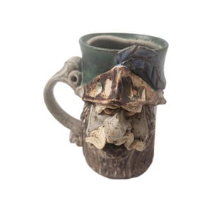May include: A handcrafted ceramic mug with a whimsical face design. The mug features a green rim, a detailed face with a beard, and a uniquely shaped handle. The color palette includes shades of brown, beige, and green.