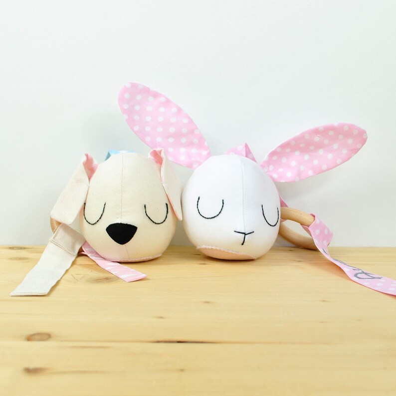 May include: A white bunny and a beige dog plush toy with embroidered faces and pink polka dot ears. The bunny has a wooden ring attached to its ear. The toys are on a light brown wooden surface.