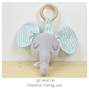 May include: A gray elephant head rattle with green and white chevron patterned ears. The rattle has a wooden ring attached to the top.