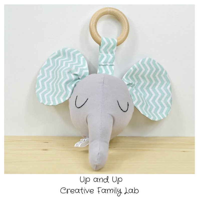 May include: A gray elephant head rattle with green and white chevron patterned ears. The rattle has a wooden ring attached to the top.