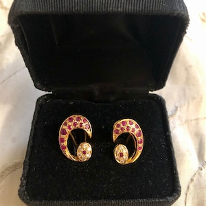 May include: A pair of gold earrings with a crescent moon design. The earrings are set with small red rubies and white diamonds.