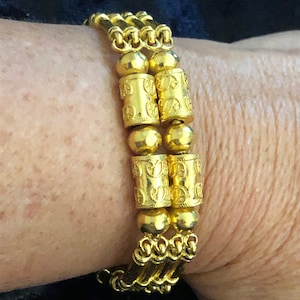 Puede incluir: Un brazalete de oro con un dise&ntilde;o de cadena triple y cuentas cil&iacute;ndricas decorativas. Las cuentas tienen un patr&oacute;n repetitivo de peque&ntilde;os c&iacute;rculos.