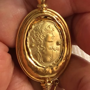 Op de afbeelding: Ovale broche van verguld metaal met een reliëfbeeld van een vrouwenprofiel. De broche heeft een paarse edelsteen bovenop en een gouden ketting.