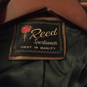 Op de afbeelding: Een bruin leren jack met een zwart label waarop "Reed Sportswear First in Quality" staat. Het label heeft een rood en goud logo met een gestileerde "R" erin.