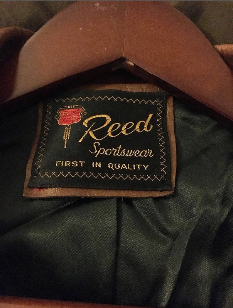 Op de afbeelding: Een bruin leren jack met een zwart label waarop "Reed Sportswear First in Quality" staat. Het label heeft een rood en goud logo met een gestileerde "R" erin.