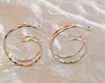 May include: A pair of silver hoop earrings with a textured, wavy design. The earrings are double-hooped, creating a layered effect.