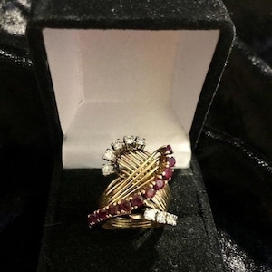 Op de afbeelding: Een gouden ring met een ontwerp van verweven draden, bezet met rode robijnen en diamanten. De ring ligt in een zwarte fluwelen doos.