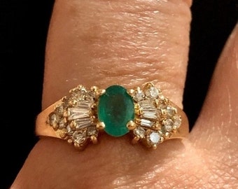 Peut inclure: Une bague en or avec une grande émeraude ovale de couleur verte entourée de diamants plus petits. La bague est portée sur un doigt.