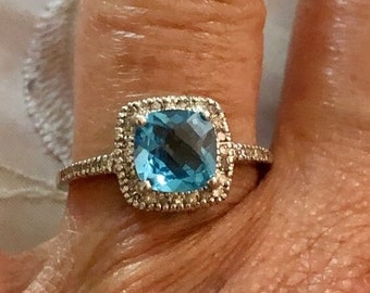 May include: A silver ring with a large, square, blue topaz gemstone surrounded by a halo of smaller diamonds.