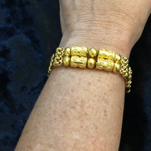 Puede incluir: Un brazalete de oro con una doble fila de eslabones de cadena y cuentas cil&iacute;ndricas decorativas. Las cuentas tienen dise&ntilde;os intrincados y est&aacute;n conectadas por cuentas de oro m&aacute;s peque&ntilde;as.
