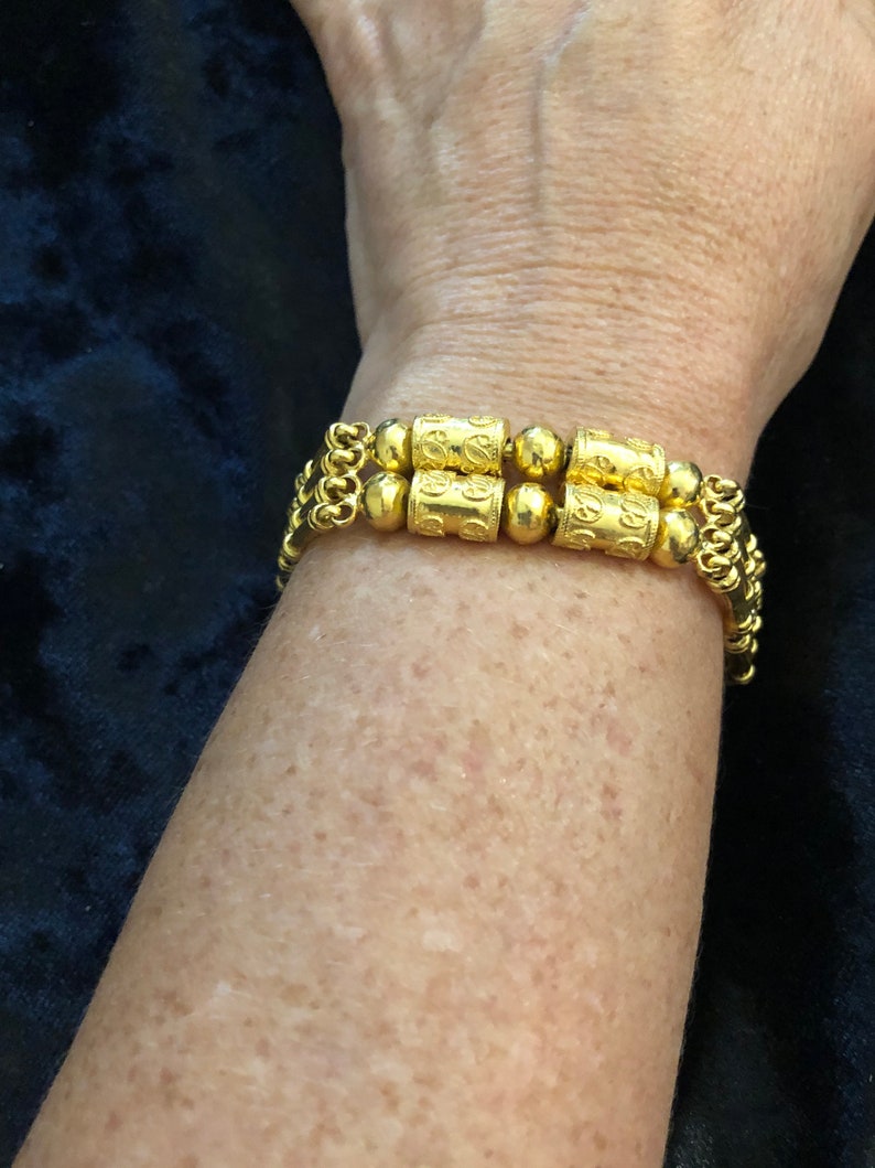 Puede incluir: Un brazalete de oro con una doble fila de eslabones de cadena y cuentas cil&iacute;ndricas decorativas. Las cuentas tienen dise&ntilde;os intrincados y est&aacute;n conectadas por cuentas de oro m&aacute;s peque&ntilde;as.