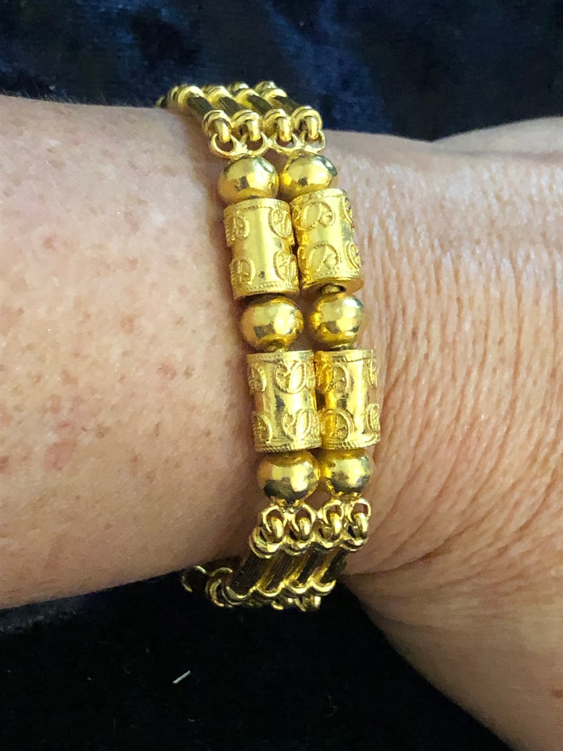 Puede incluir: Un brazalete de oro con un dise&ntilde;o de cadena triple y cuentas cil&iacute;ndricas decorativas. Las cuentas tienen un patr&oacute;n repetitivo de peque&ntilde;os c&iacute;rculos.