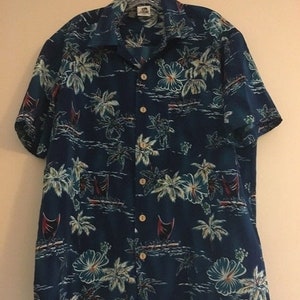 Op de afbeelding: Een blauw, kort mouwig overhemd met knopen en een tropische print met palmbomen, bloemen en zeilboten. Het overhemd heeft een witte kraag en knopen.