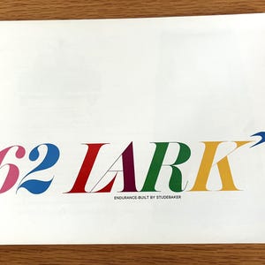 May include: A white paper with the colorful text "'62 LARK" in pink, blue, red, purple, green, and yellow. The text "ENDURANCE-BUILT BY STUDEBAKER" is below the main text. A blue arrow graphic is on the right.