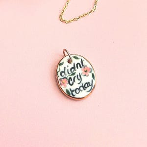 Peut inclure: Un collier à chaîne dorée avec un pendentif ovale en céramique portant l'inscription peinte à la main "didn't cry today" entourée de petites fleurs roses et de feuilles vertes, accompagné d'une boîte cadeau en carton brun avec une étiquette.