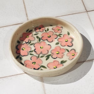 May include: A small, round ceramic dish with a cream-coloured base. The dish is decorated with pink flowers and green leaves. The flowers have a simple, hand-painted style. The dish is likely used for serving small portions of food or as a decorative item.