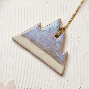 May include: A ceramic Christmas tree decoration with a light blue and cream glaze. The tree-shaped ornament has a gold string for hanging. The ornament is on a white, textured surface.