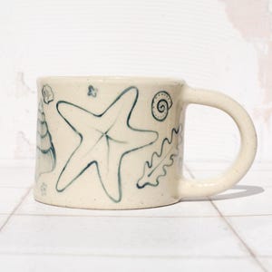 May include: Cream-colored ceramic mug with hand-drawn blue sea life illustrations. The design includes a starfish, seashells, and seaweed. The mug has a rounded handle and a simple, clean design, perfect for enjoying a warm beverage.