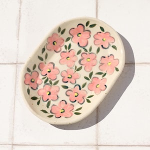May include: Oval ceramic dish with a cream base, decorated with hand-painted pink flowers, green leaves, and yellow centres. The floral design is evenly spaced. The dish is on a white tiled surface.