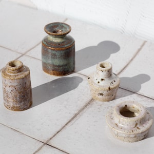 May include: Four small ceramic vessels with varied shapes and textures. One is a tall, mottled brown and green cylinder, another is a textured beige cylinder, and two are shorter, wider pieces in speckled off-white with flared rims and openings.