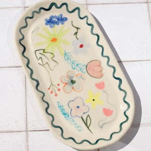 May include: Oval ceramic serving tray with a cream base and a wavy, dark teal border. The tray is decorated with colourful, whimsical floral designs in shades of pink, yellow, blue, and green.