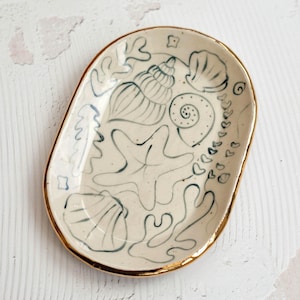 Peut inclure: Plat ovale en céramique avec une base crème et un bord doré. Le plat est décoré d'illustrations bleues dessinées à la main de coquillages, d'étoiles de mer et d'algues. Le plat mesure environ 15 cm de long et 10 cm de large.