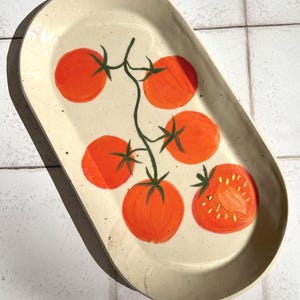 May include: An oval-shaped ceramic platter with a cream-coloured speckled glaze. The centre features a hand-painted design of a green vine with five whole red tomatoes and one halved tomato showing yellow seeds, rendered in a vibrant orange-red hue.
