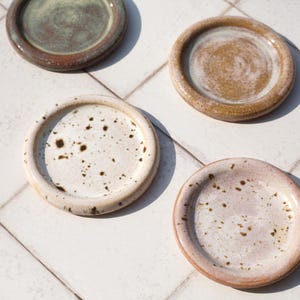 May include: Four circular ceramic coasters with raised edges. One is dark green and brown, one is tan, one is off-white with dark brown speckles, and one is light pink with dark brown speckles. Each coaster has a smooth, glazed finish.